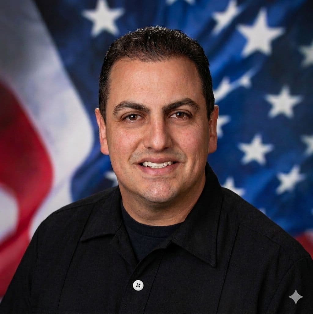 Smiling man in a black shirt with a blurred American flag in the background.